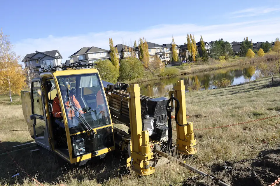 A yellow directional drill in the ground next to a community pond.